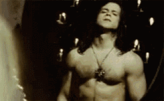a shirtless man with long hair and a necklace is standing in a dark room .