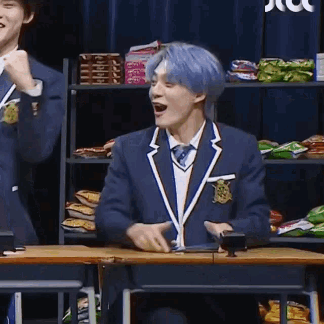 a man with blue hair is sitting at a desk in front of a shelf of snacks .