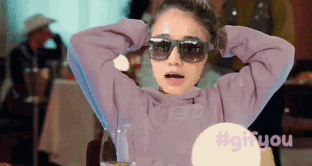 a woman wearing sunglasses and a purple sweatshirt is sitting at a table with her hands behind her head .