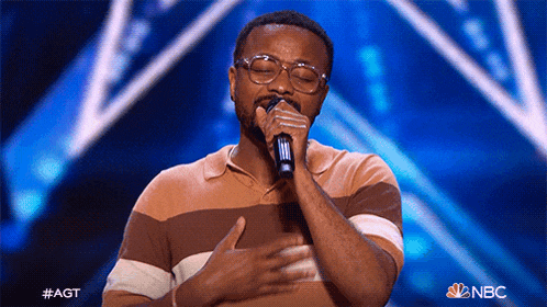 a man wearing glasses is singing into a microphone on a stage with a nbc logo in the background