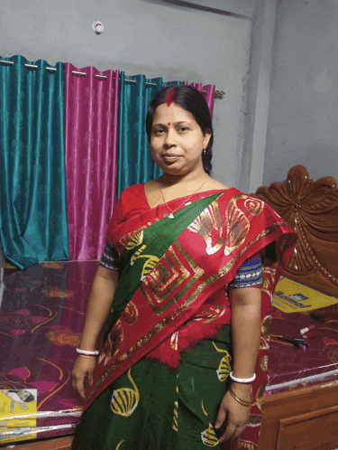 a woman in a red and green saree stands in front of a bed with a mattress on it