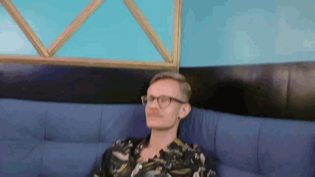 a man wearing glasses sits on a blue couch in front of a window