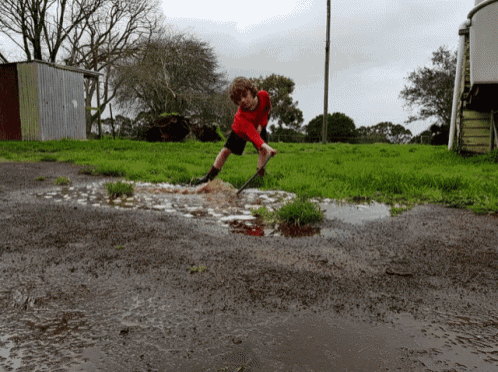 a boy in a red shirt is playing in a muddy puddle with a shovel