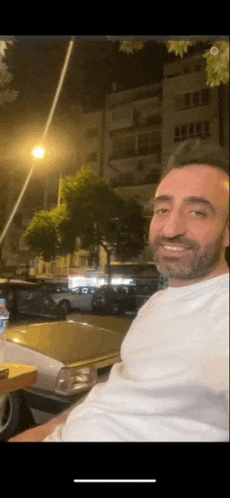 a man with a beard is smiling while sitting at a table in front of a car .