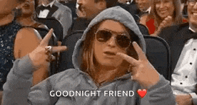 a woman in a hoodie and sunglasses is sitting in a crowd of people and giving a peace sign .