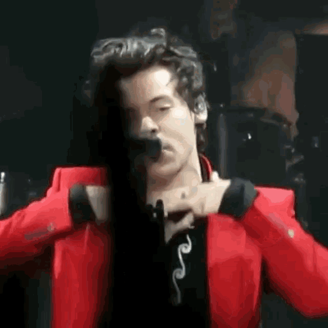 harry styles is wearing a red jacket and smoking a cigarette while singing into a microphone on stage .