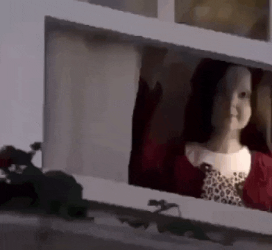 a doll is looking out of a window and waving at the camera .