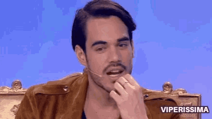 a man with a mustache is pointing at his face while sitting in a chair .