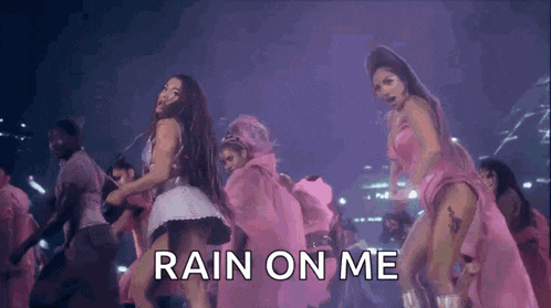 a group of women in pink dresses are dancing on a stage with the words rain on me above them .