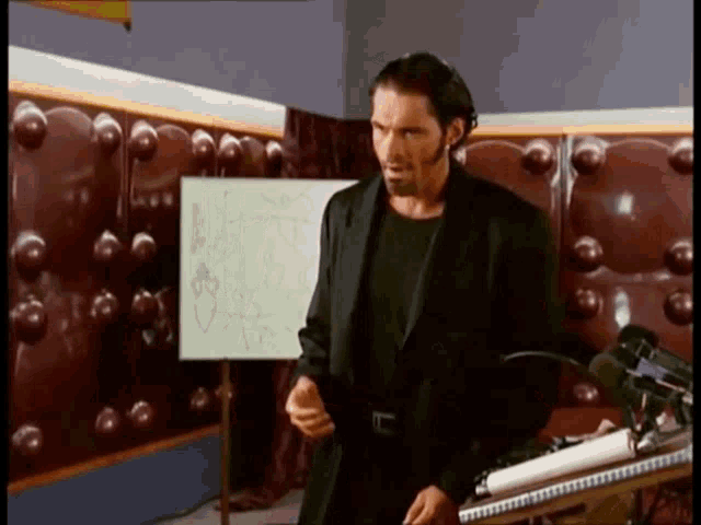 a man in a black suit stands in front of a white board with a drawing of a bird on it