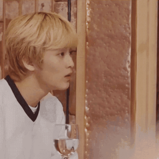 a young man with blonde hair is sitting at a table with a glass of water and a sign that says what on it .