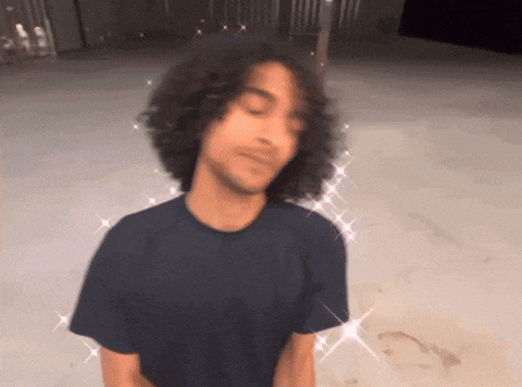 a man with curly hair wearing a black t-shirt is standing in a parking lot .