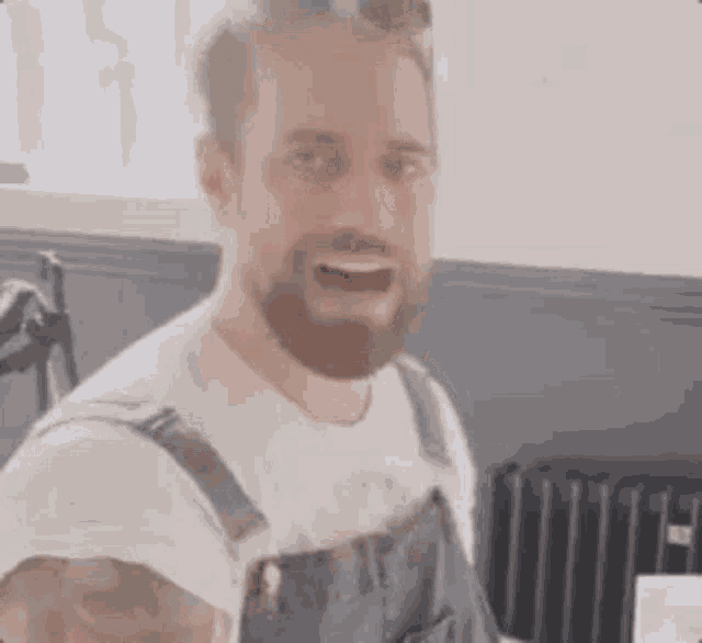 a man with a beard is wearing overalls and a white shirt and is making a funny face .