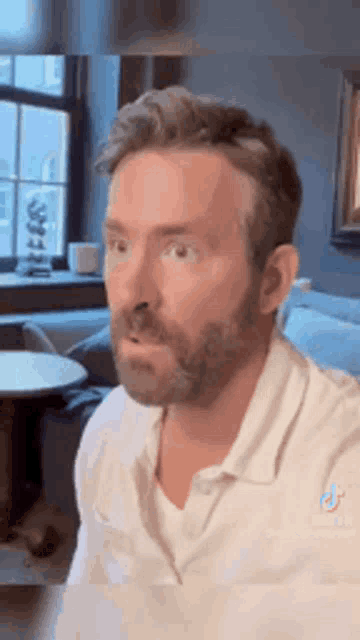 a man with a beard and a white shirt is sitting in a living room with his mouth open .