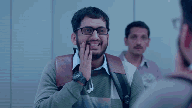 a man wearing glasses and ear buds smiles with another man behind him