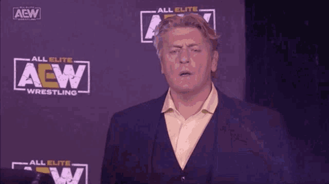a man in a suit and tie is standing in front of a purple wall with a aew logo on it .