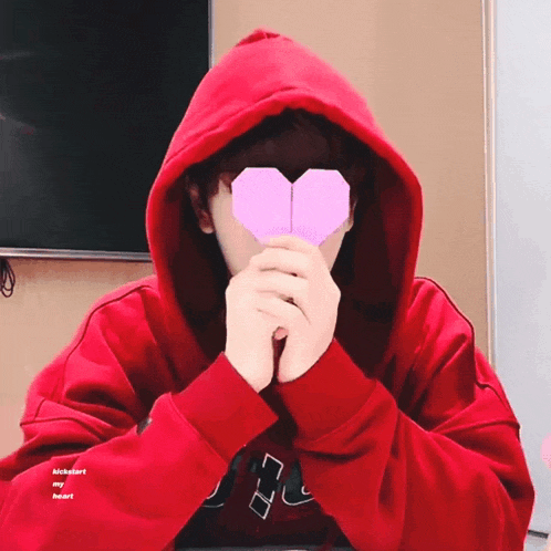 a person wearing a red hoodie is holding an origami heart in front of their face ..