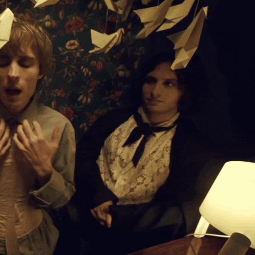 two men are sitting next to each other in a room with origami birds hanging from the ceiling