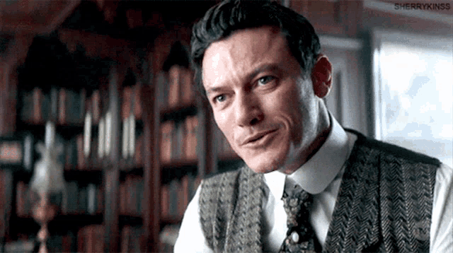 a man in a vest and tie is smiling in front of a bookshelf .