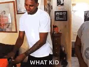 a man in a white shirt and black shorts is standing in a living room holding a dog and says `` what kid '' .