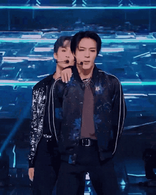 a couple of men are standing on a stage . one of the men is wearing a bomber jacket .