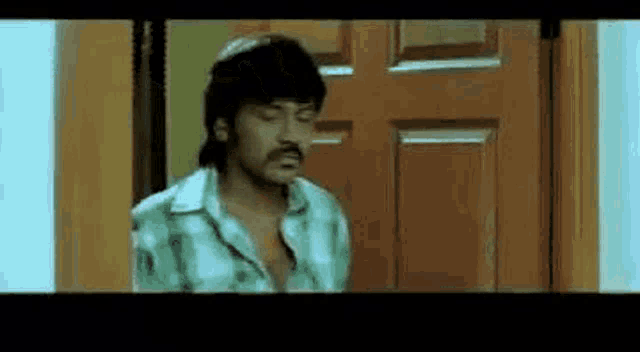 a man with a mustache is standing in front of a door and looking at the camera .