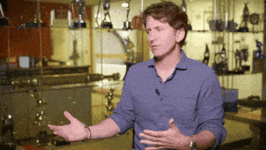 a man in a blue shirt is talking in front of trophies on a shelf