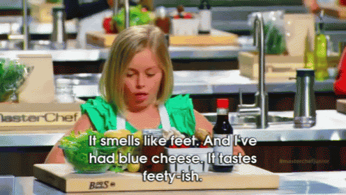 a little girl in a green apron sits at a cutting board with a bowl of food and a bottle of sauce