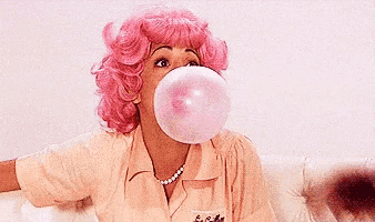 a woman in a pink wig is blowing a bubble of gum while sitting on a couch .