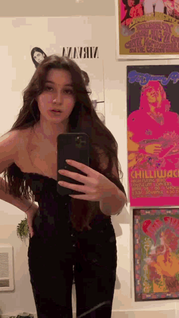 a woman is taking a selfie in front of a mirror with posters on the wall behind her .