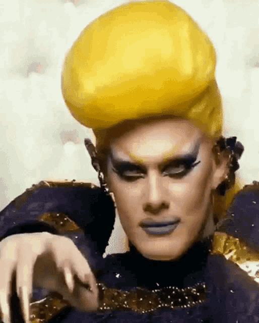 a drag queen with long nails and a yellow wig is wearing a purple and gold costume .