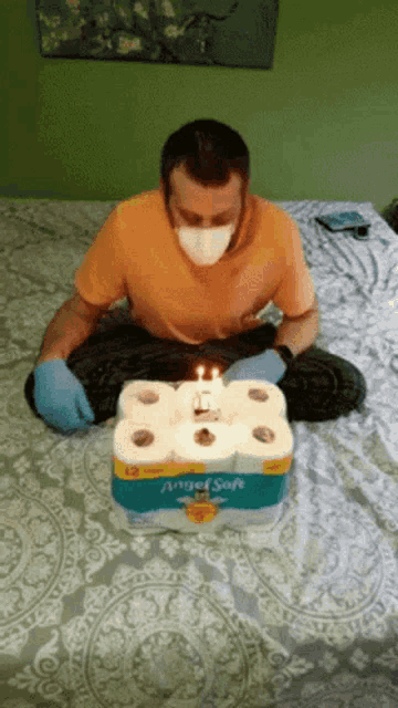 a man wearing a mask and gloves is sitting on a bed next to a box of angel soft toilet paper