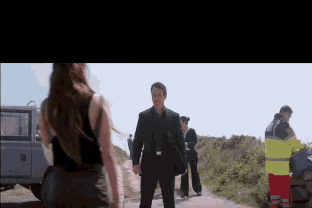 a man in a suit and a woman in a black tank top are walking down a road