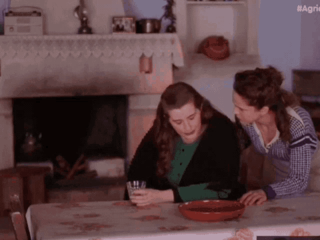 two women are sitting at a table and one is holding a glass while the other looks on