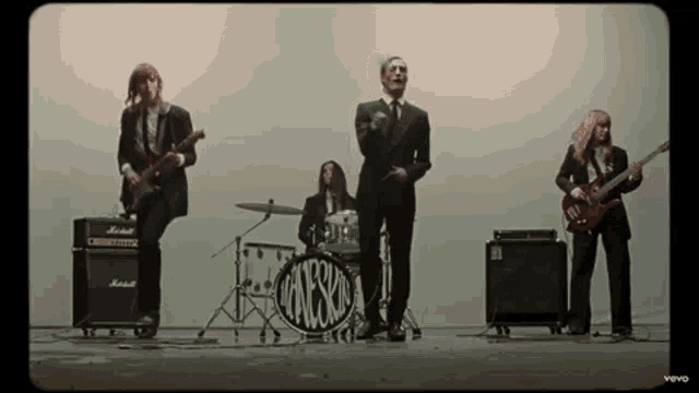 a group of men in suits are standing on a stage playing guitars and drums .