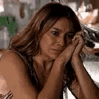 a woman is sitting at a table with her hands on her chin and looking at the camera .