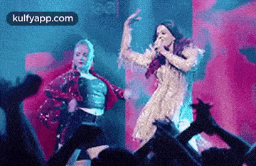 a woman is singing into a microphone while another woman is dancing in front of a crowd at a concert .