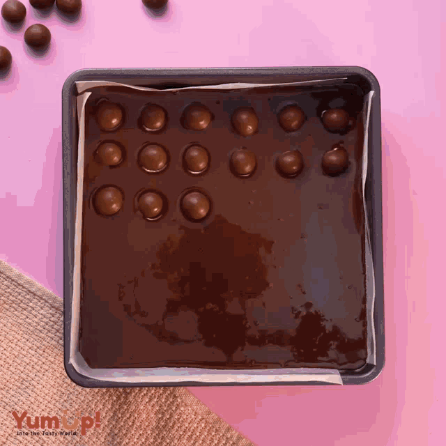 a square pan filled with chocolate and maltesers on a table with a pink background that says yumop