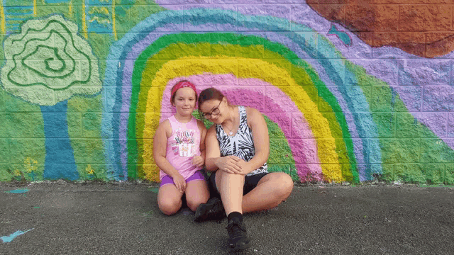 two girls sitting in front of a rainbow mural