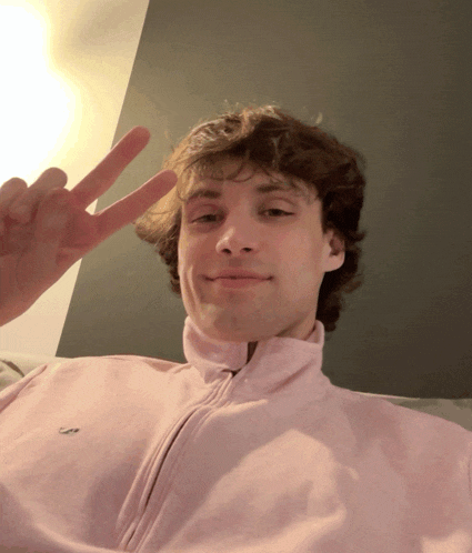 a young man wearing a pink lacoste sweatshirt giving a peace sign