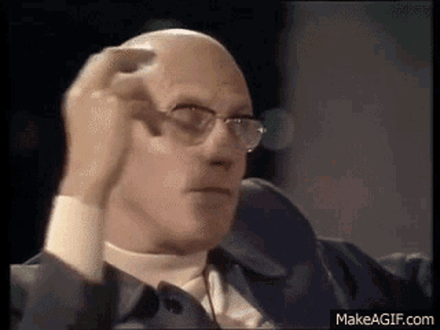 a bald man wearing glasses and a suit is scratching his head with his hand .