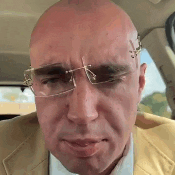 a bald man wearing glasses and a suit is sitting in a car with his eyes closed .