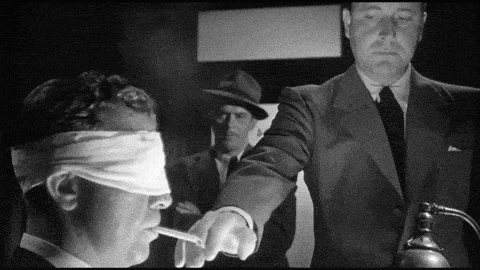 a man with blindfolds on his eyes is smoking a cigarette while another man points at him