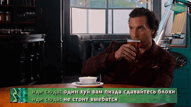 a man sitting at a table drinking a glass of beer with a green sign above him that says " иди сюда "