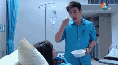 a man in a blue shirt is feeding a woman in a hospital bed with a spoon