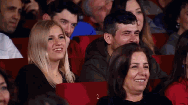 a group of people are sitting in red seats in a theater with the number 2 on the bottom right