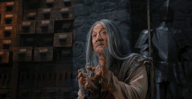 a man with long grey hair and a beard is holding something in his hands in front of a wall with drawers and a knight