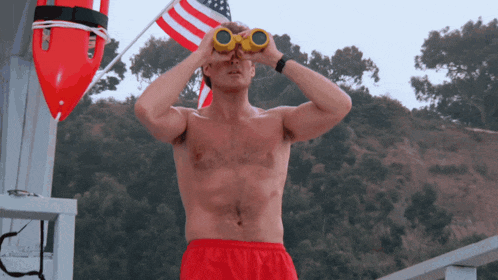 a shirtless man looks through binoculars in front of a life preserver
