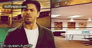 a man in a suit and white shirt is standing in a room with a sign that says srk-queen.tumblr .