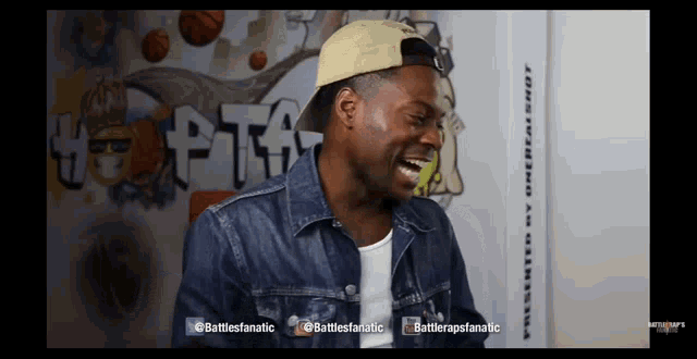 a man wearing a hat and a denim jacket laughs in front of a wall that says battlerapfanatic